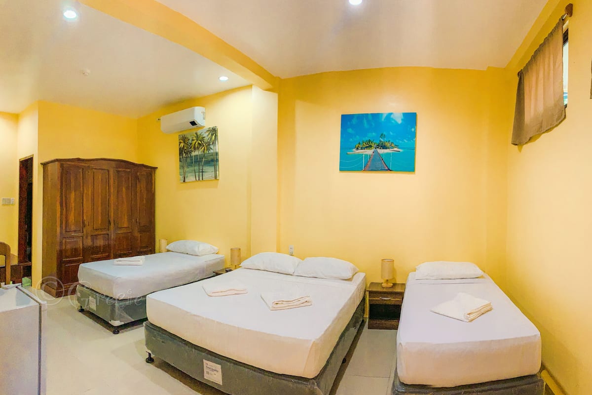 A hotel room with three single beds, a wooden wardrobe, a painting of a tropical scene, and an air conditioning unit.