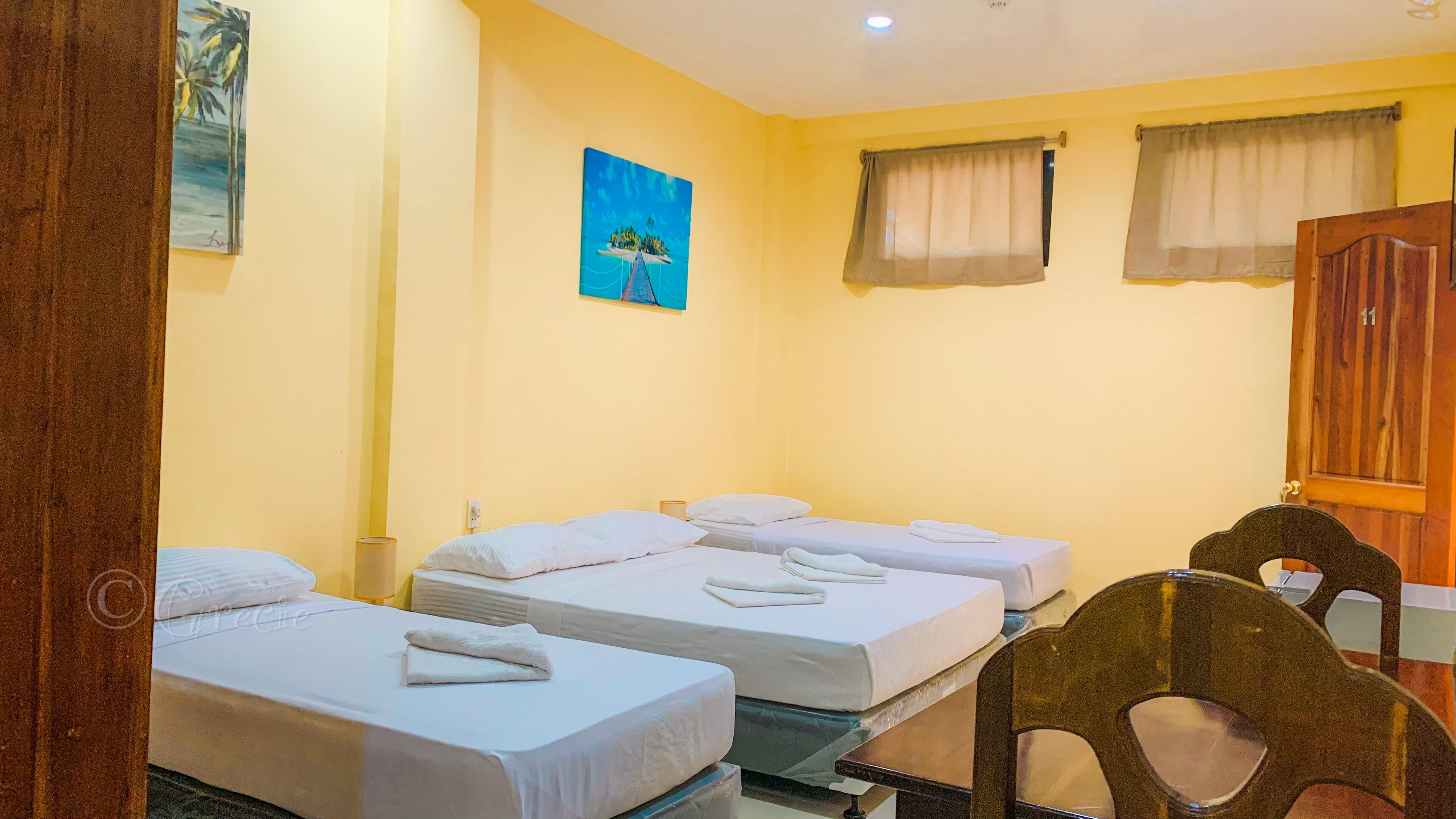 Une chambre d’hôtel avec trois lits simples, une chaise en bois, un tableau accroché au mur et une porte en bois portant un numéro.