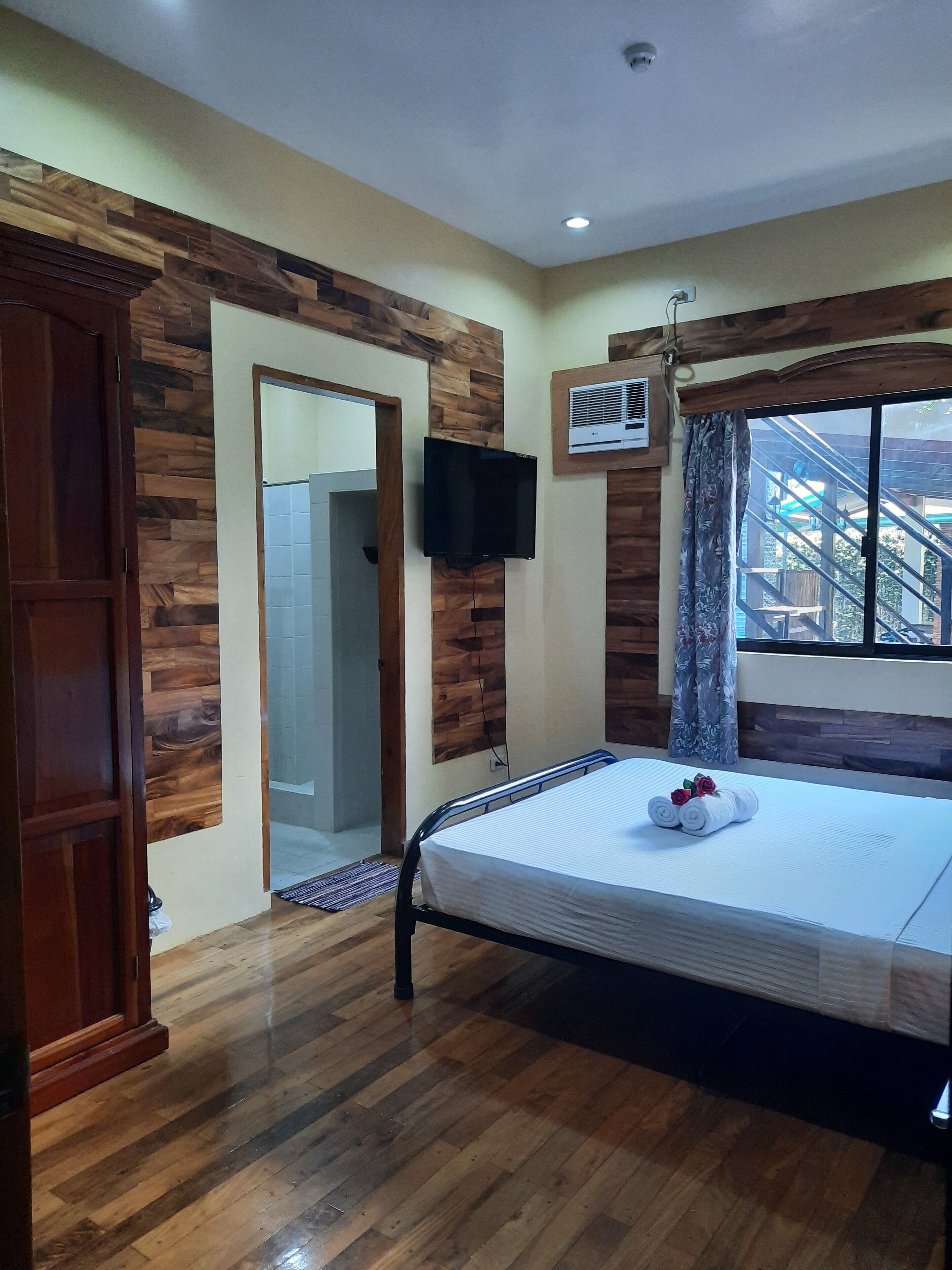 A hotel room with a wooden floor, a bed with a white mattress and rolled towels, a television mounted on the wall, and a window with curtains.