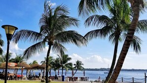 A beachfront area with palm trees, a thatched-roof bar, and outdoor seating by the water. - Picturesque View Suites (Johor Bahru)