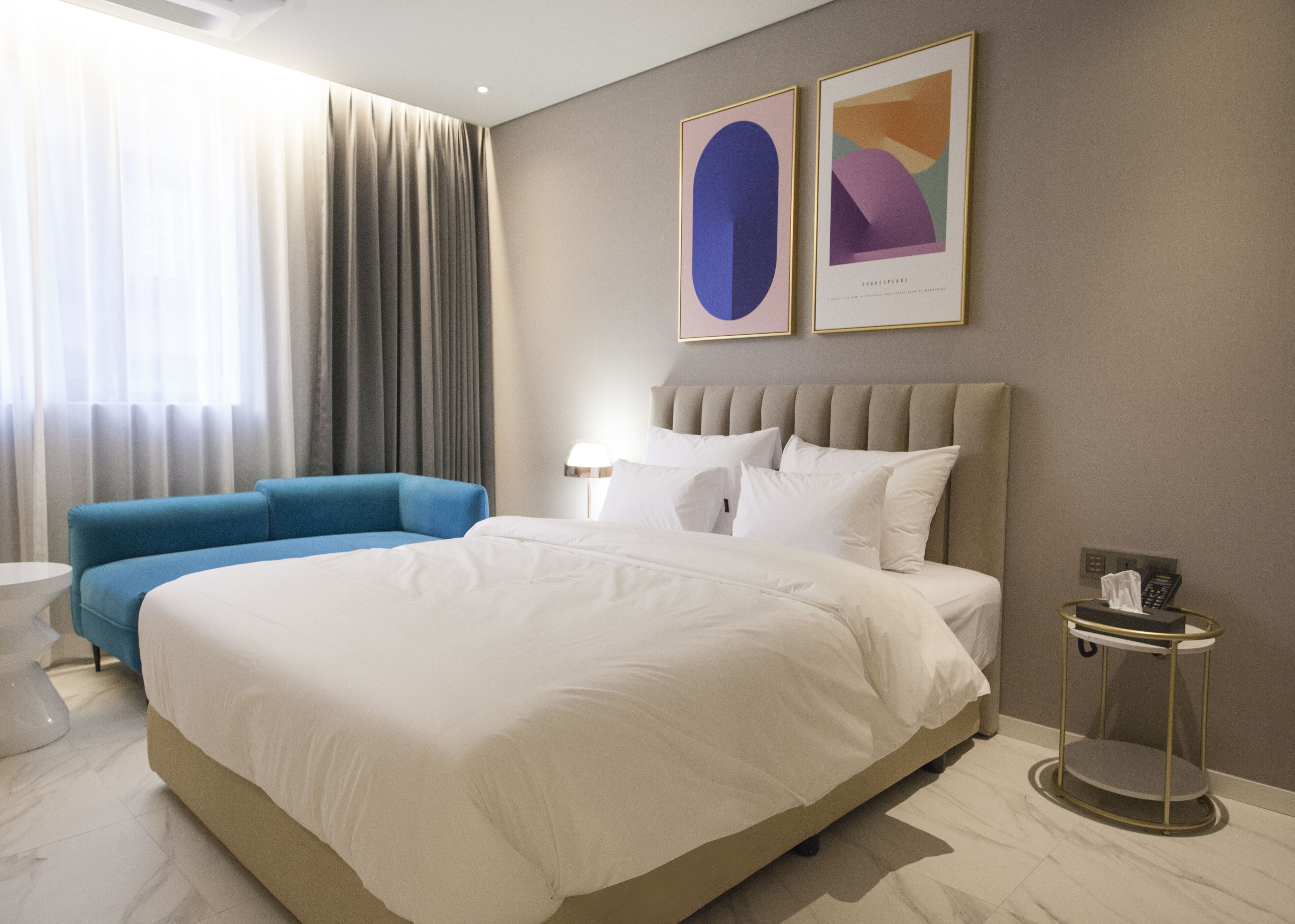 A modern hotel room with a large bed, a blue sofa, a bedside table, and two framed artworks on the wall.