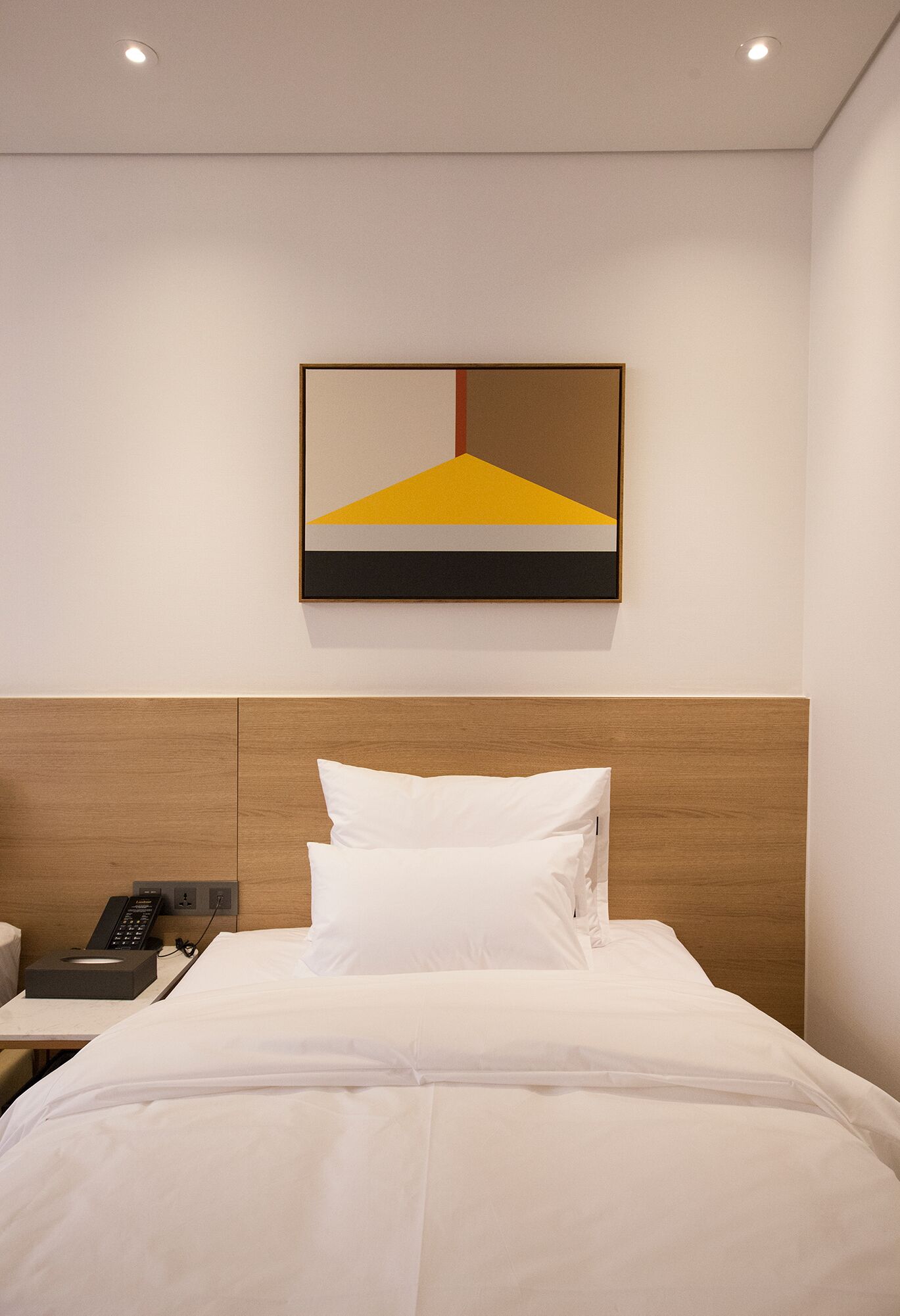 A hotel room with a wooden headboard, a bed with white bedding, a bedside table with a phone, and a painting on the wall.