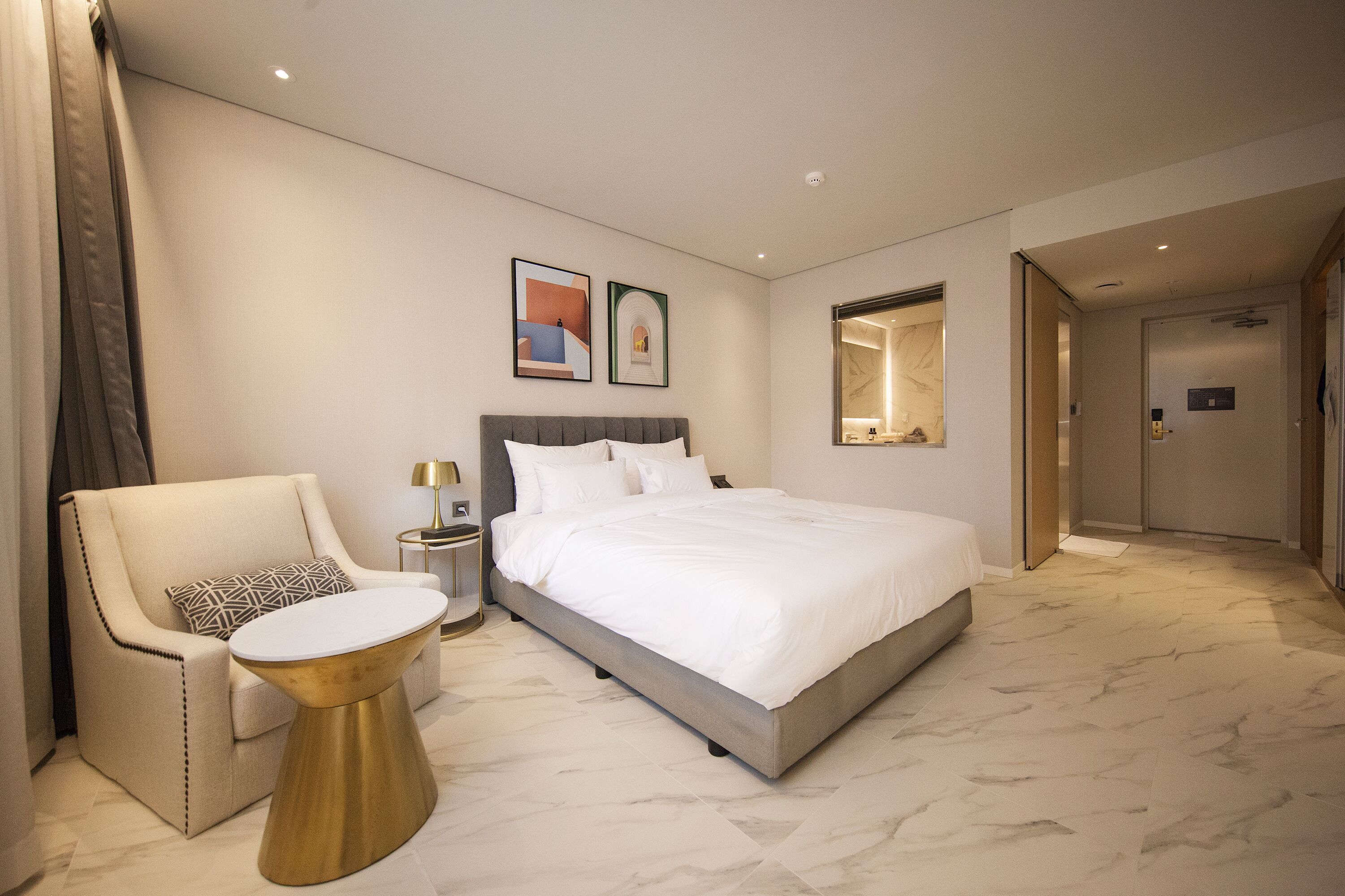 A modern hotel room with a large bed, a sitting area with a chair and a small table, and a mirror on the wall.