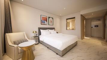 A modern hotel room with a large bed, a sitting area with a chair and a small table, and a mirror on the wall.