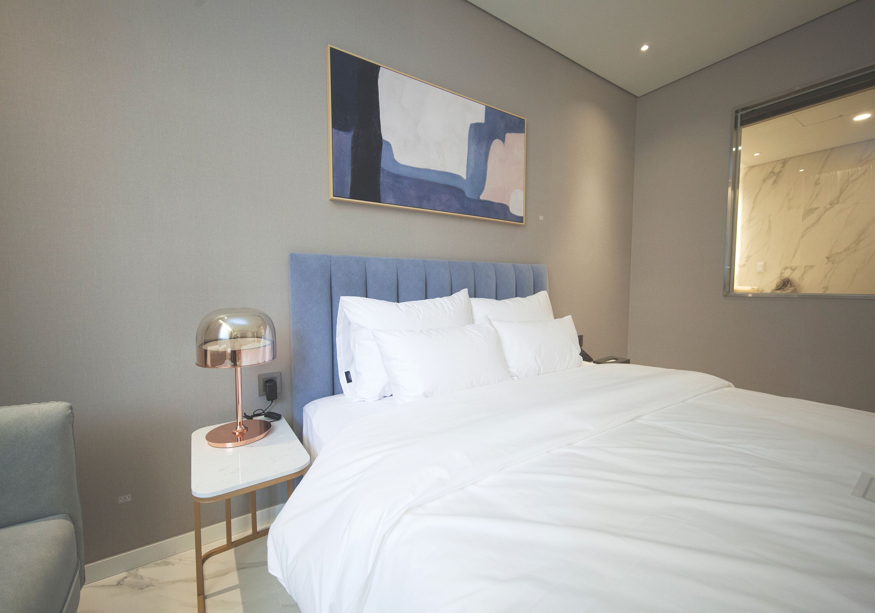 A modern hotel room with a large bed, a bedside table with a lamp, a chair, and a painting on the wall.