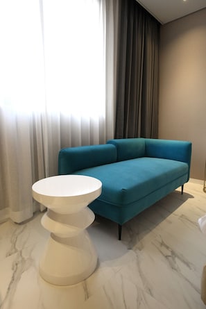 A modern living room with a teal sofa, a white round coffee table, and a large window with sheer curtains.