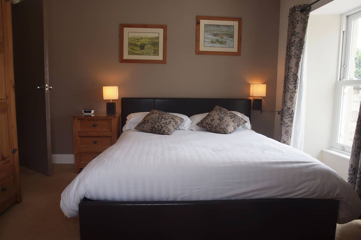 A bedroom with a large bed, two bedside lamps, a nightstand with a radio, and two framed pictures on the wall.
