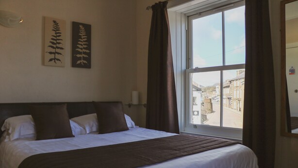 A neatly made bed with a brown blanket, two wall-mounted fern paintings, a window with curtains, and a view of buildings outside. - Herriot's in Hawes (Hawes)