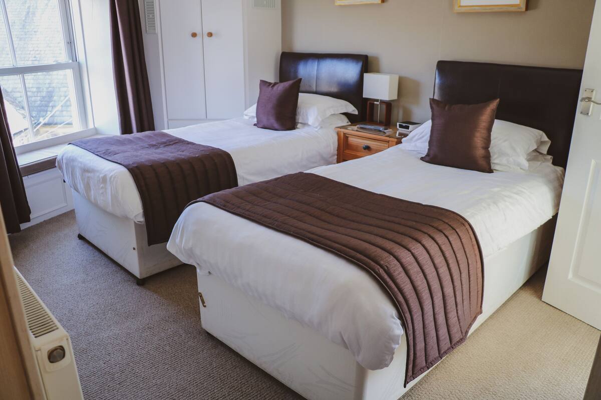 A hotel room with two beds, each with a brown blanket and pillows, a bedside table with a lamp, and a window with curtains.