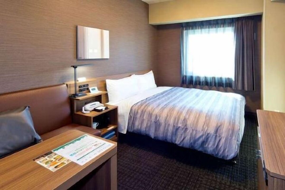 Een hotelkamer met een bed, nachtkastje, lamp en een raam met gordijnen.