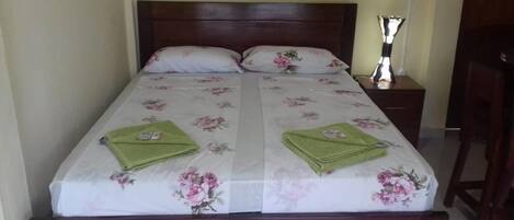A double bed with floral bedding, two pillows, a bedside table with a lamp, and a wooden nightstand.