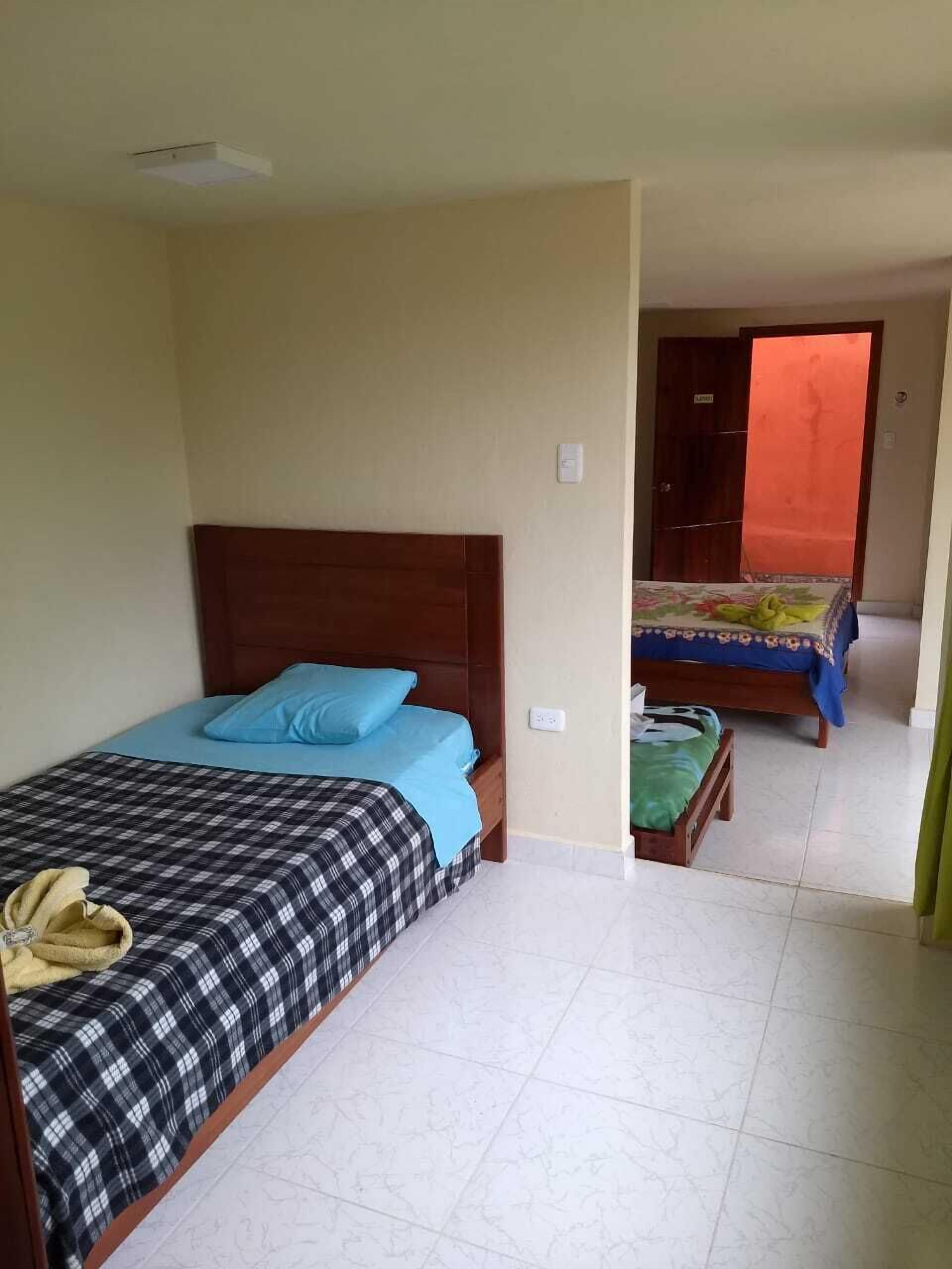 A room with two beds, one with a blue and black checkered bedspread and the other with a floral bedspread. There is a wooden headboard on the left bed, and a small wooden bench with a green cushion in front of the second bed.