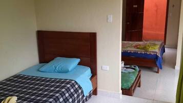 Una habitación con dos camas, una con colcha azul y negra a cuadros y la otra con estampado floral. Una cabecera de madera adorna la cama de la izquierda, y un pequeño banco de madera con cojín verde se encuentra frente a la segunda cama.