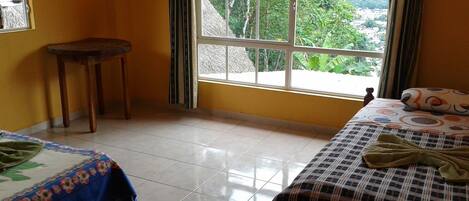 Una habitación con dos camas, una mesita y un ventanal con vistas a zonas verdes y edificios.
