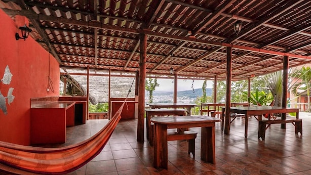 A covered outdoor seating area with wooden tables and benches, a hammock, and a red wall. - Hostal Tena Ñaui (Tena)