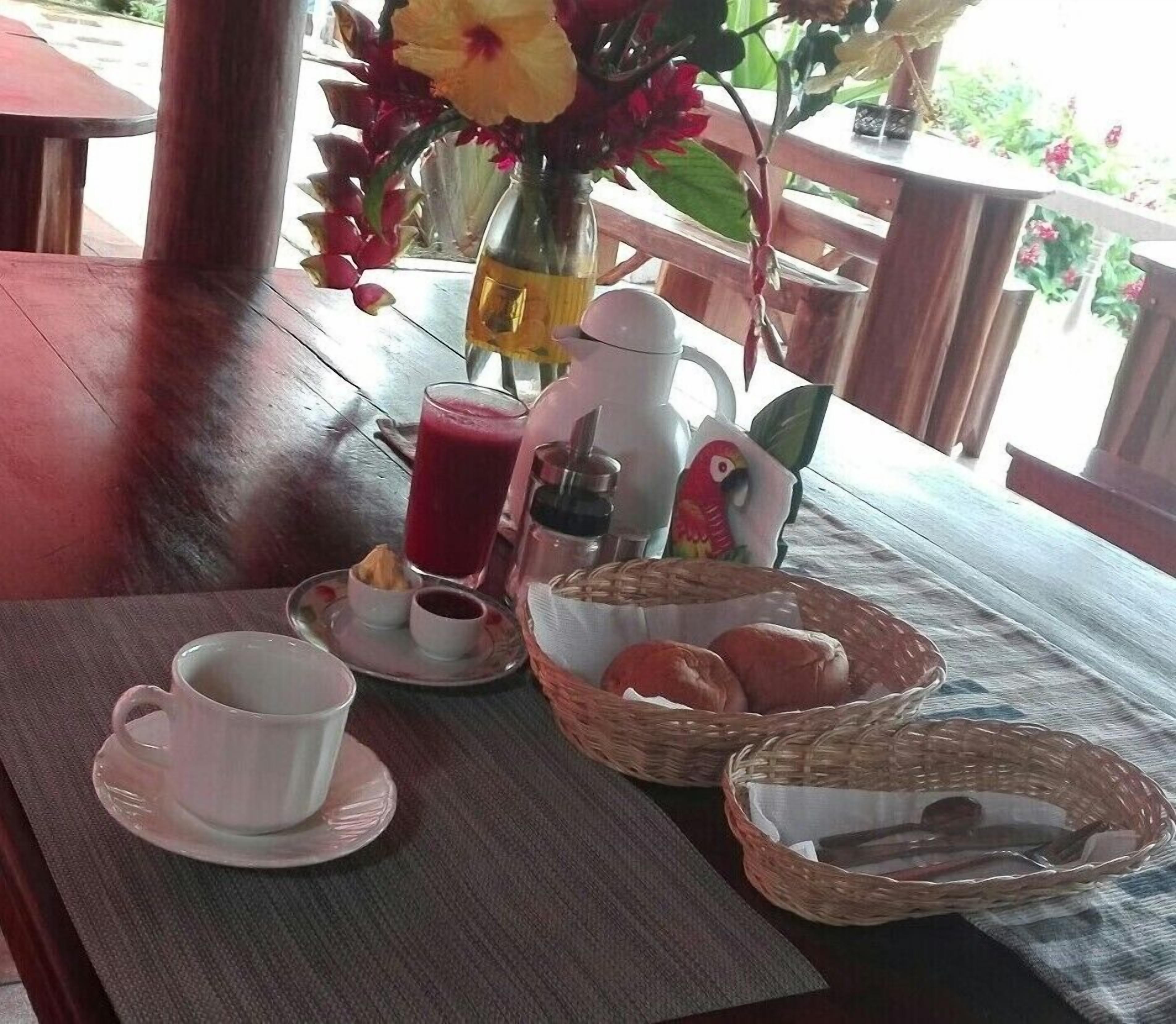 A breakfast table with a coffee maker, a pitcher, a glass of juice, a cup, a plate with pastries, and a basket of bread.