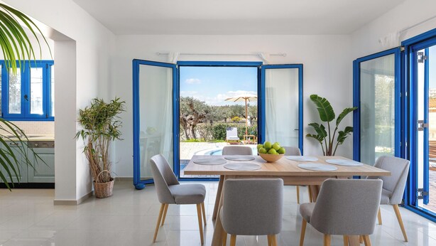 A modern dining area with a table set for four, large windows with blue frames, and potted plants. - Blue Moon Villa in Kolympia (RHODES)