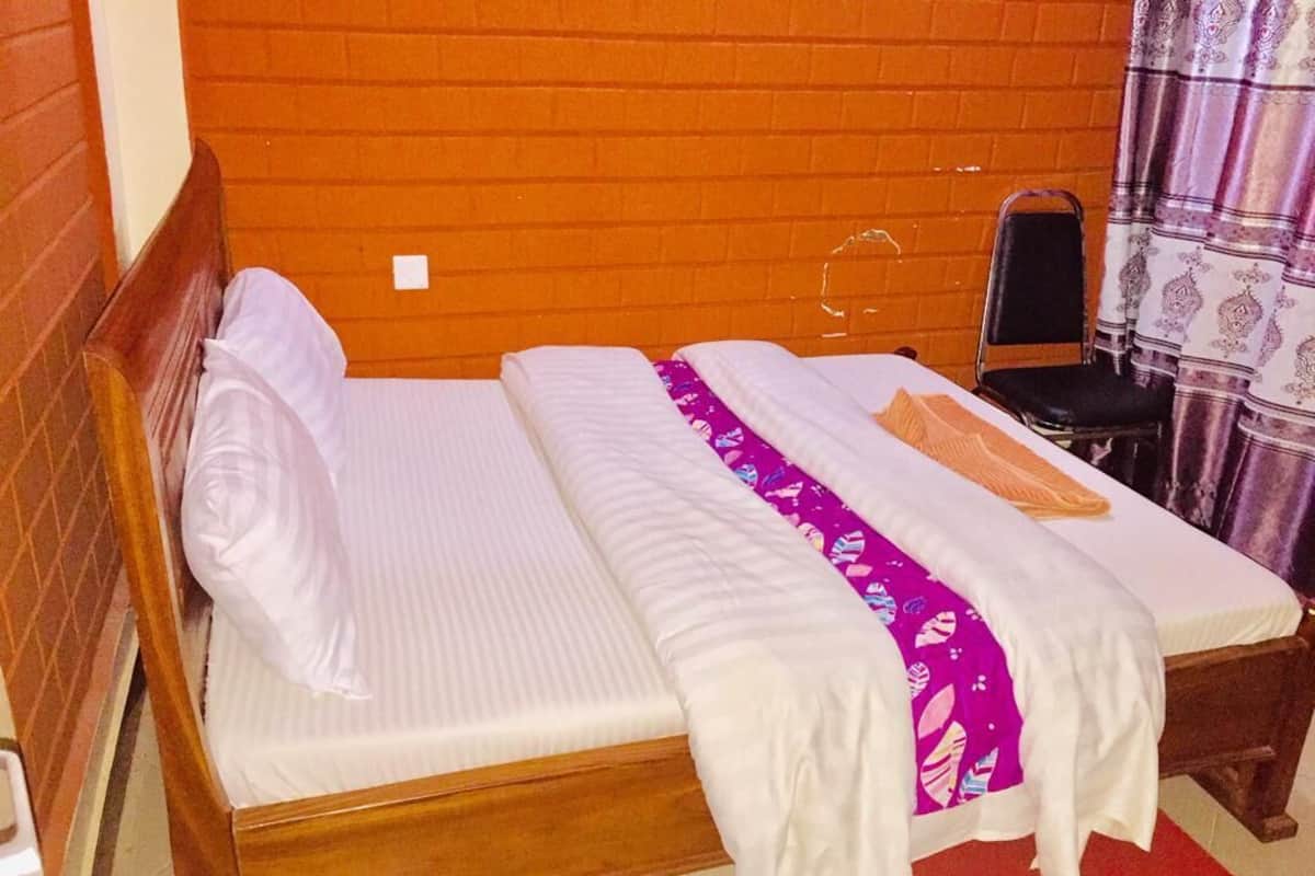 Una cama individual con cabecera de madera, colchón blanco y colcha morada. Hay una silla con asiento negro y una cortina estampada al fondo.