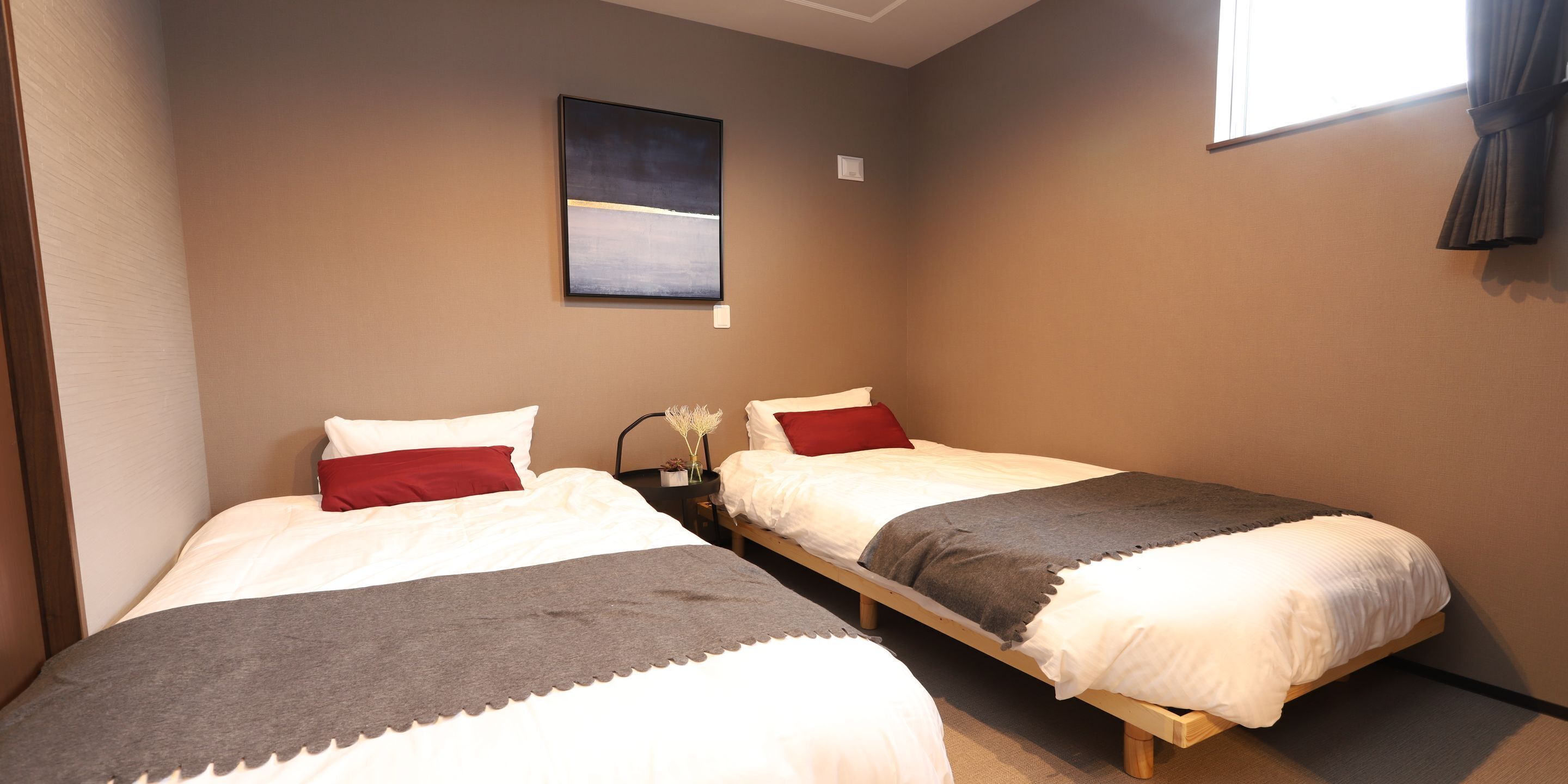 A room with two single beds, each with white bedding and red pillows. There is a painting on the wall above the beds, and a window with curtains to the right.