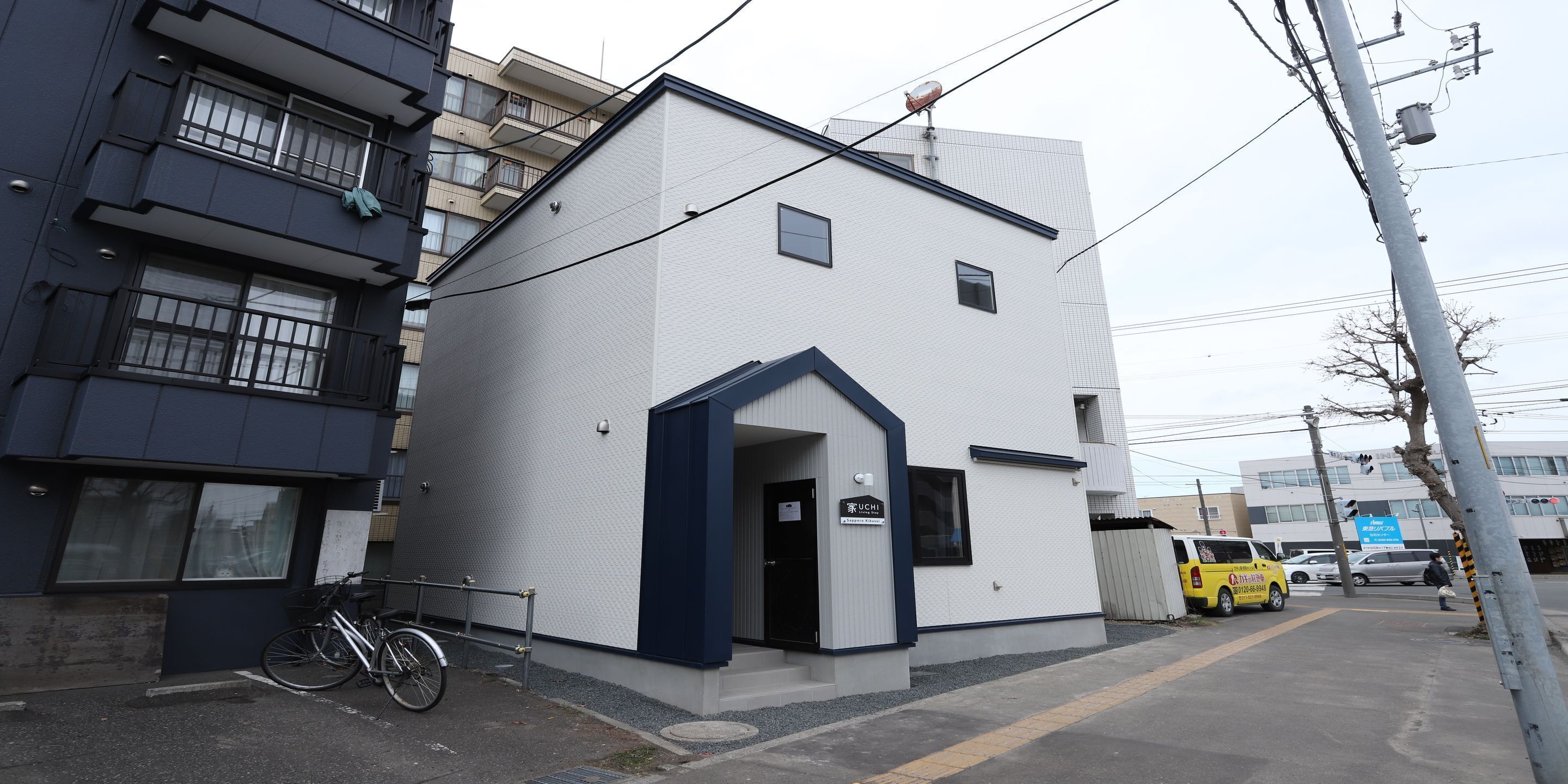Un edificio moderno con fachada blanca y detalles oscuros. Tiene una pequeña entrada con un letrero sobre la puerta. Una bicicleta está estacionada afuera y se ve una furgoneta amarilla al fondo.