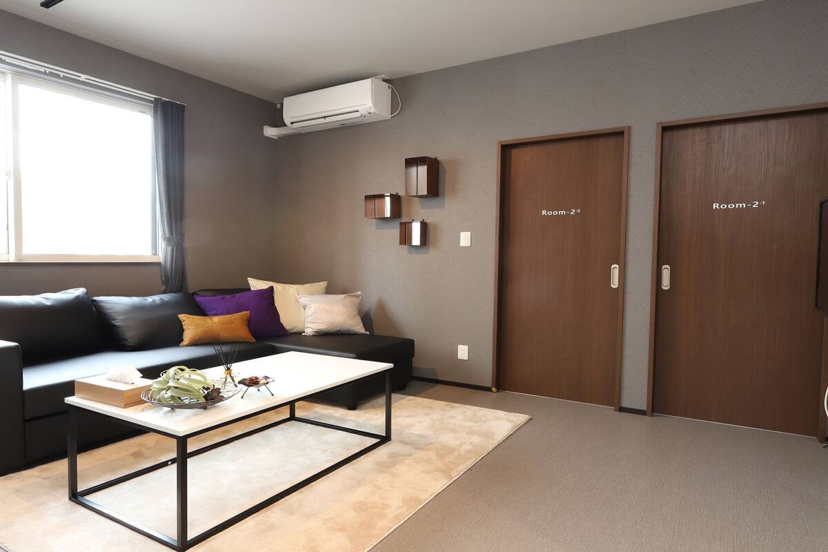 A modern living room with a black leather sofa, a white coffee table, and a beige rug. There are two wooden doors labeled "Room-2-1" and "Room-2-2".
