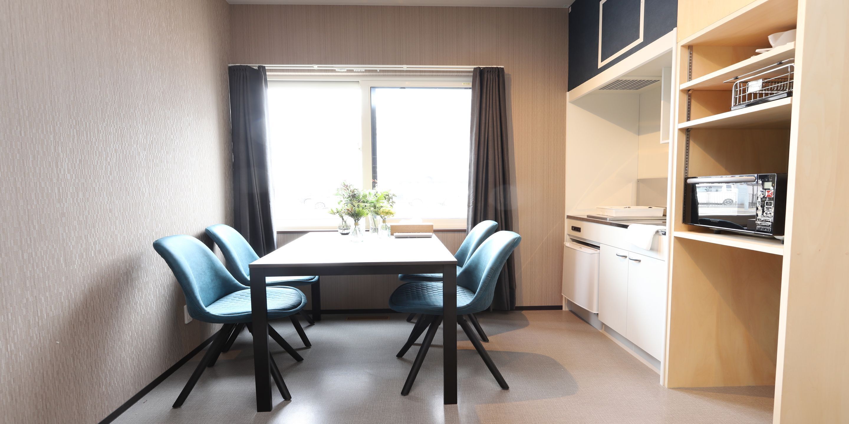 A dining area with a table, four chairs, a window with curtains, and a microwave on a shelf.