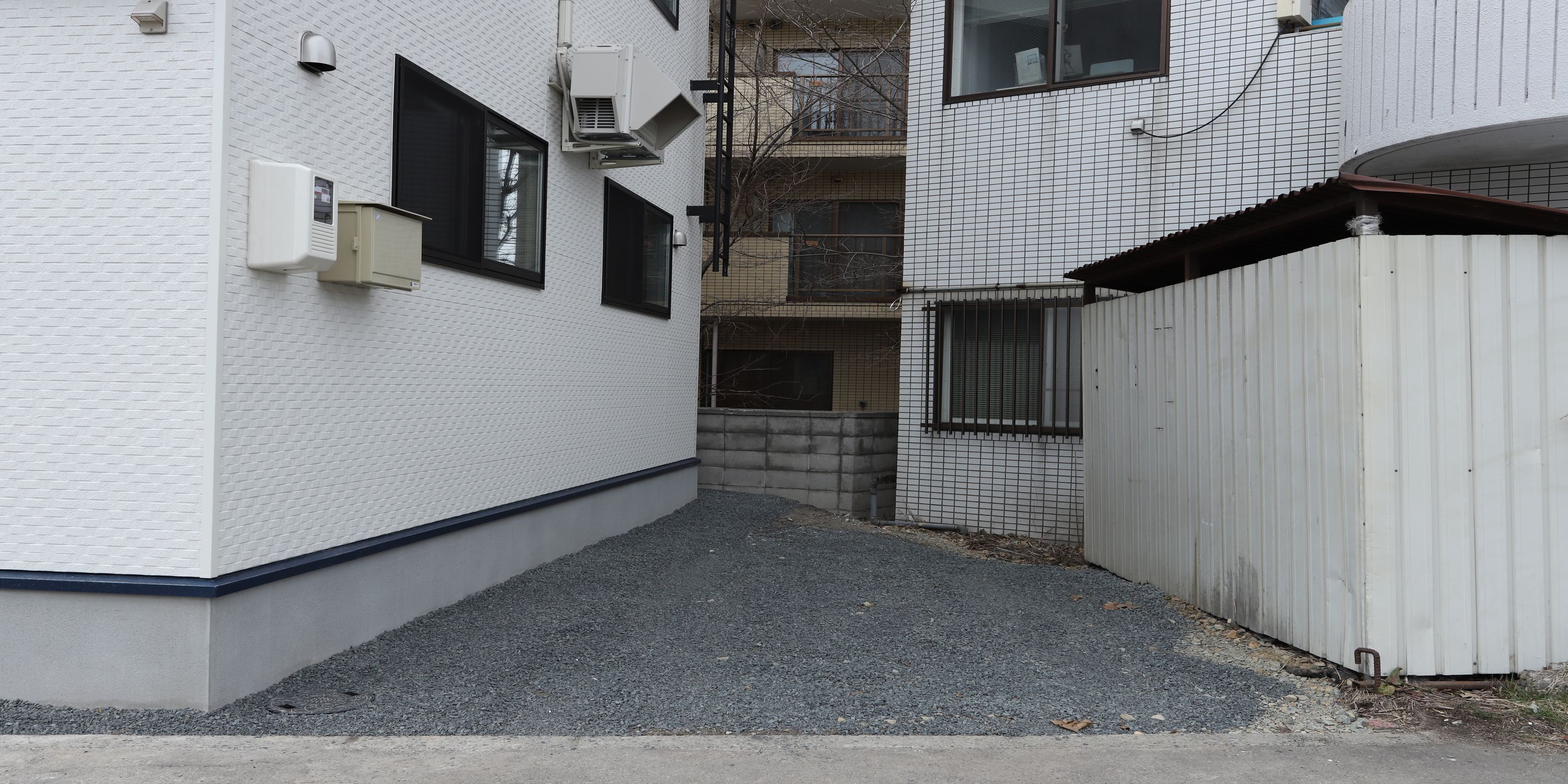 Une ruelle étroite, avec un bâtiment blanc sur la gauche, une zone délimitée par une grille sur la droite et une petite fenêtre équipée d’une caméra de surveillance.