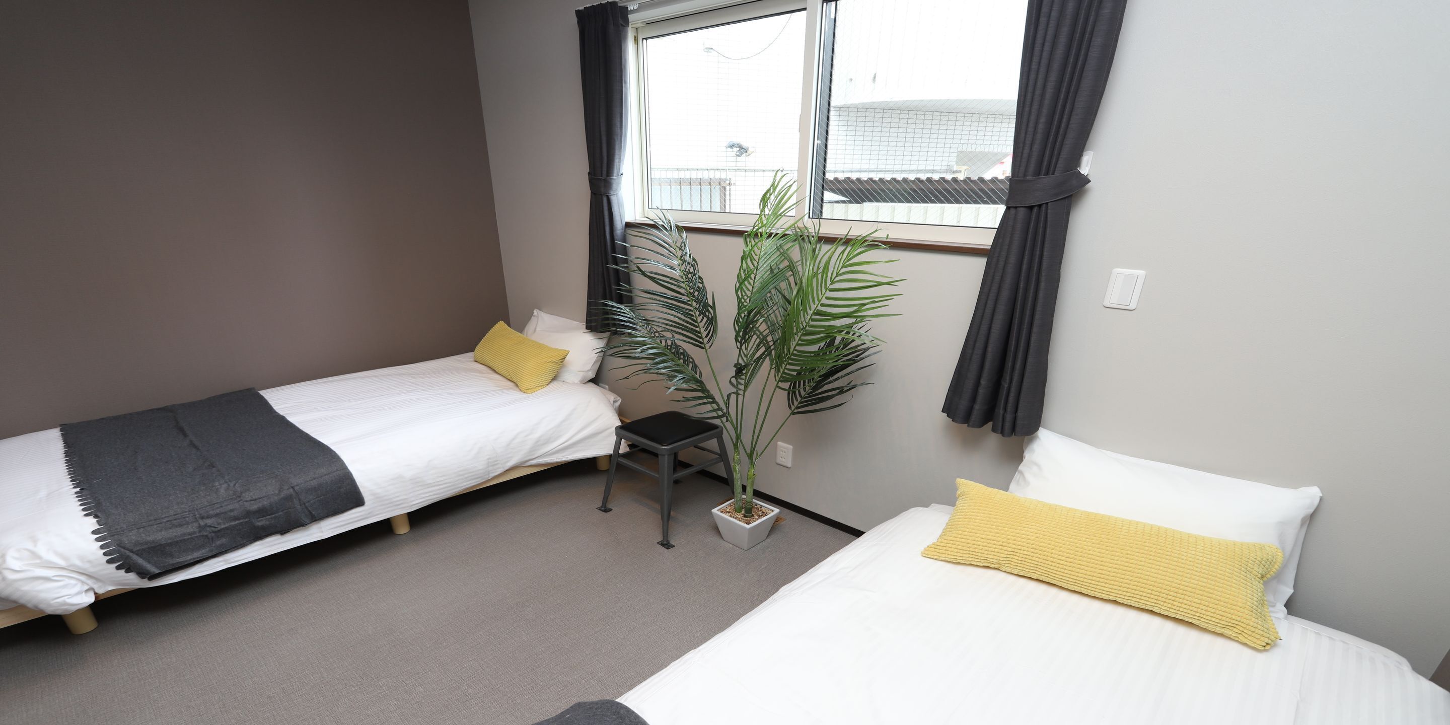 A hotel room with two single beds, a small table, a potted plant, and a window with curtains.