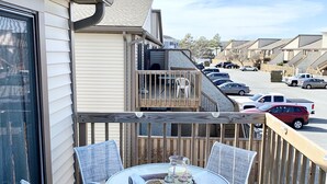 Un balcon avec une table ronde et des chaises en osier blanc, donnant sur un parking et les bâtiments voisins.