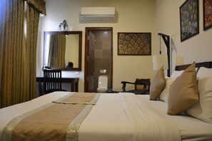 A hotel room with a large bed, a desk, a chair, a mirror, and a door. - Hotel Shambhu Villas (Udaipur)