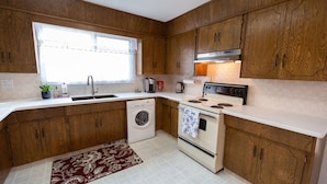 A compact kitchen with wooden cabinets, a built-in oven, a washing machine, and a window providing natural light. - Central, 2 bedroom King/Queen, Semidetached House in French Quarter (Edmonton)