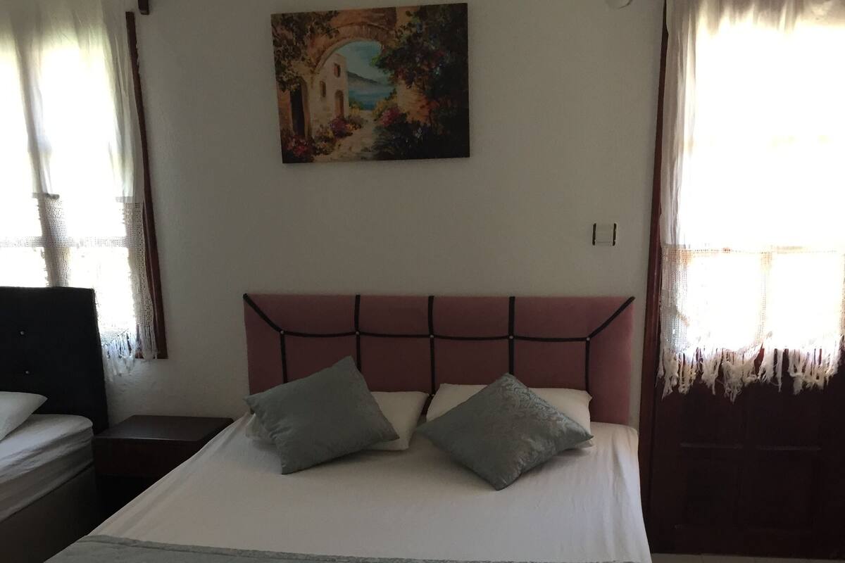 A neatly made bed with a headboard, a painting on the wall, and a window with curtains.