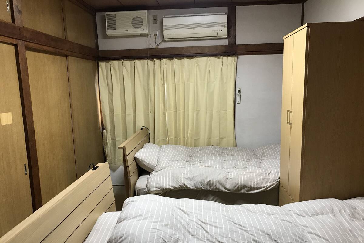A room with two single beds, wooden headboards, a window with curtains, and an air conditioning unit above the beds.