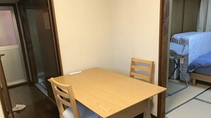 A small, wooden dining table with two chairs, a single chair with a cushion, and a bed with a blue blanket in the adjacent room. - Michio's House (Ebina)