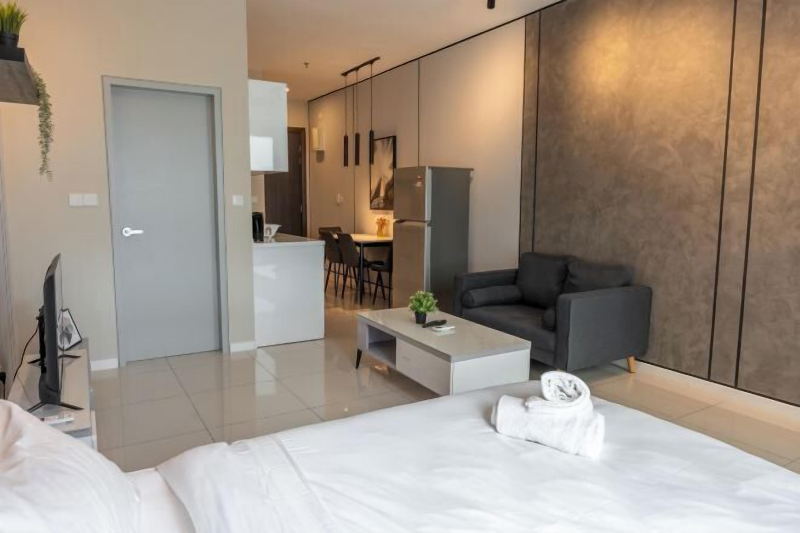 A modern hotel room with a bed, a gray sofa, a small coffee table, and a kitchenette in the background.