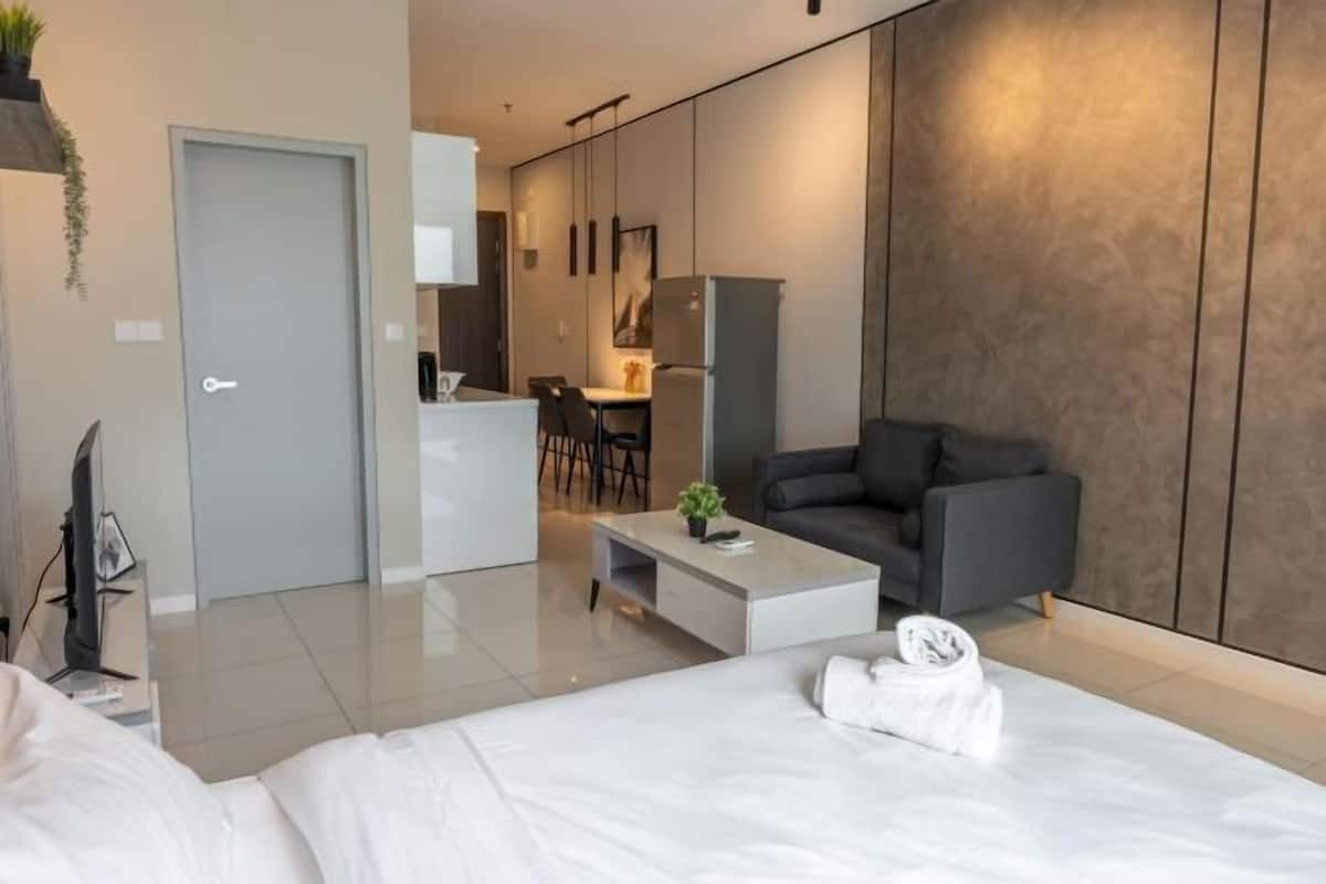 A modern hotel room with a bed, a gray sofa, a small coffee table, and a kitchenette in the background.