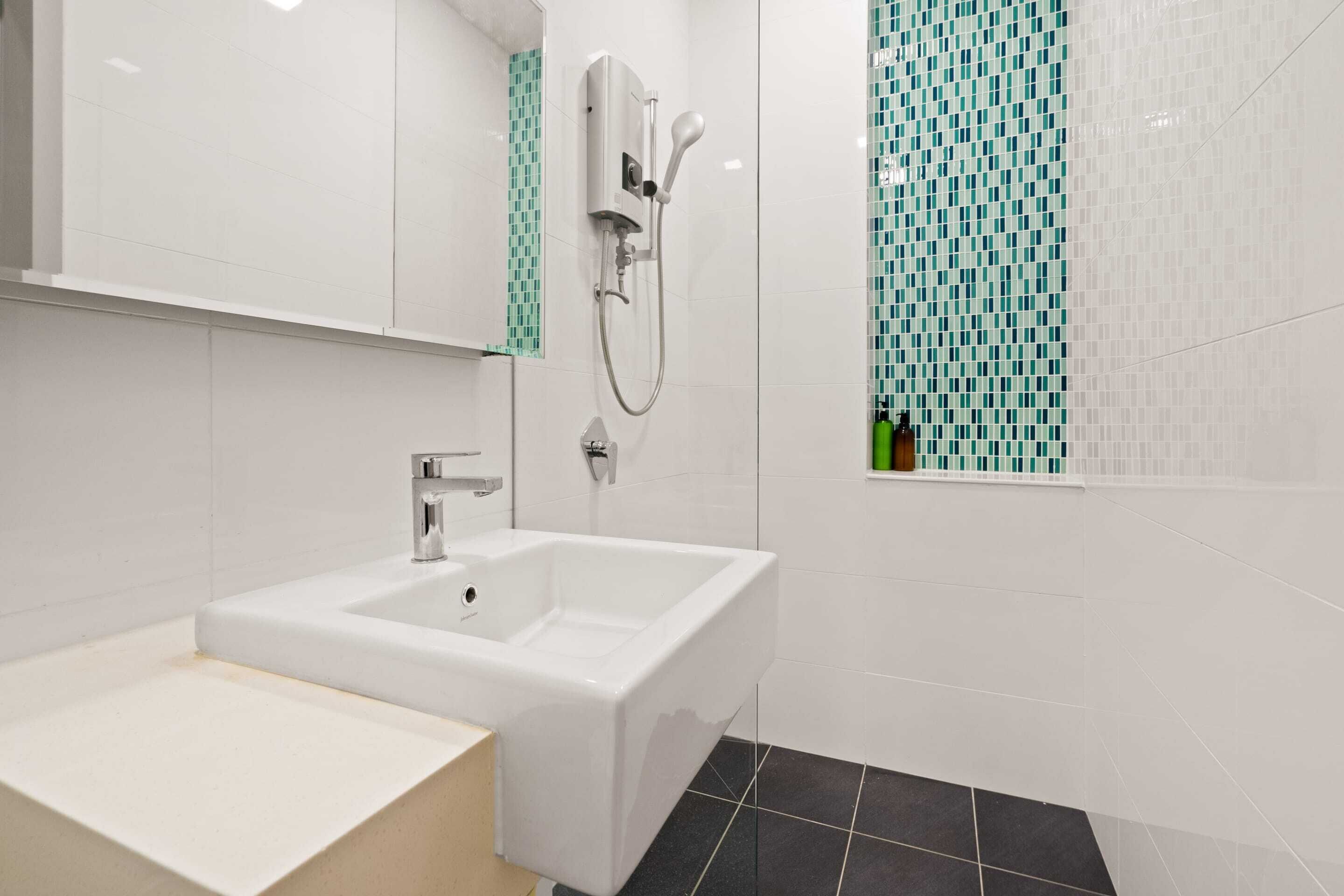 Ein modernes Badezimmer mit einem weißen Waschbecken, einer Handbrause und einer gefliesten Wand mit grünem Mosaikmuster.