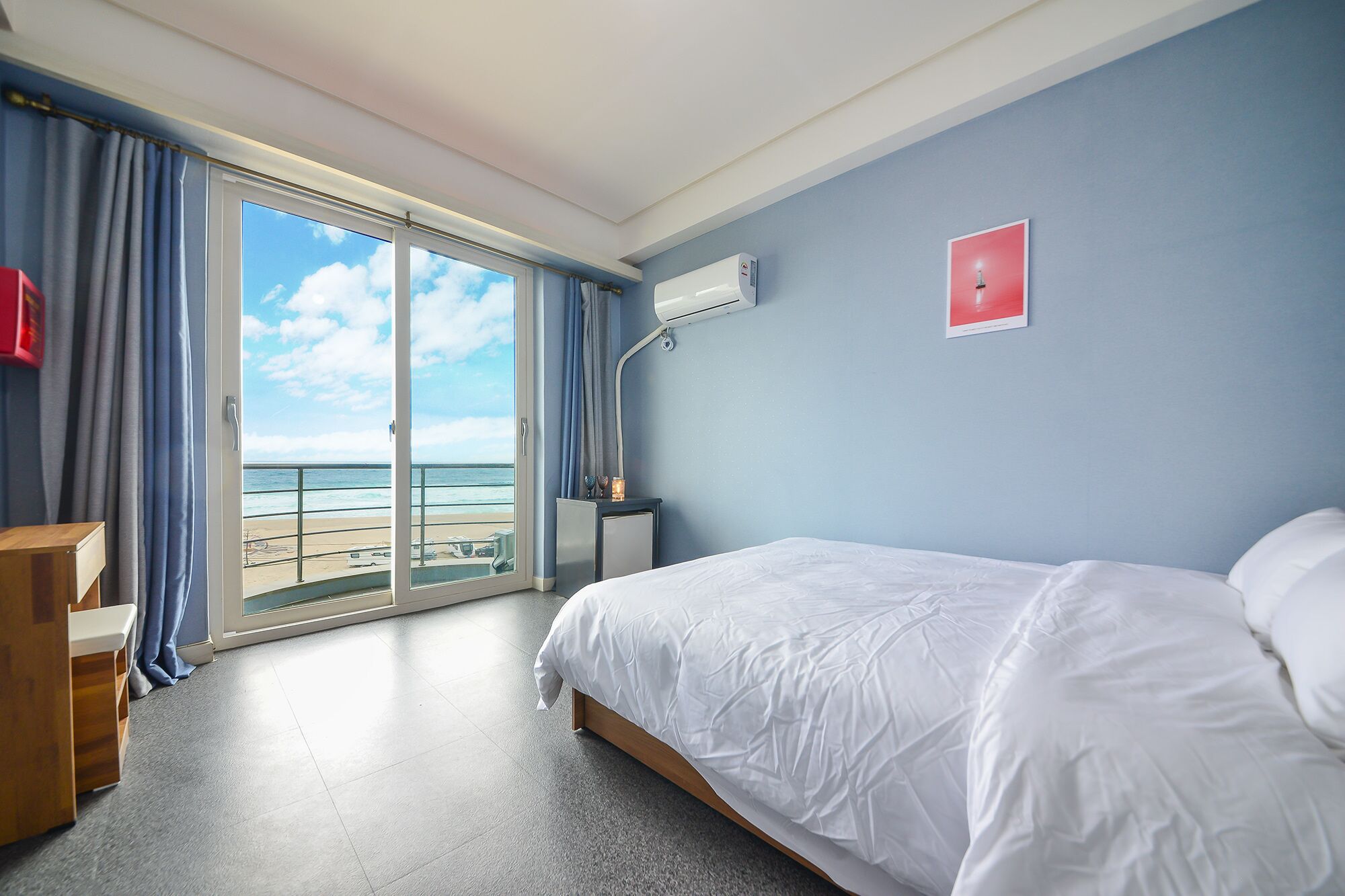 A hotel room with a bed, a desk, a chair, a window with a view of the beach, and an air conditioning unit.
