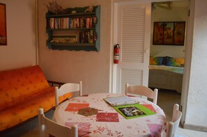 A cozy room with a round table, white chairs, a bookshelf, and a bed with colorful bedding.