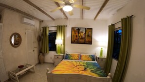 Une chambre à coucher comprenant un lit, des tables de chevet, un ventilateur de plafond, une fenêtre avec des rideaux, un climatiseur mural et un tableau.