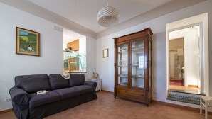 A living room with a dark grey sofa, a wooden cabinet with glass doors, a framed picture on the wall, and a view into the kitchen through an open doorway.