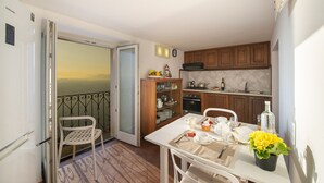 A compact kitchen with wooden cabinets, a dining table set for two, and a view of the sea through an open door.