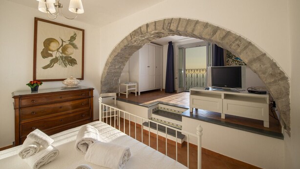 A bedroom with a stone archway, a wooden dresser, a bed with white linens, a television, and a small table with a vase of flowers.