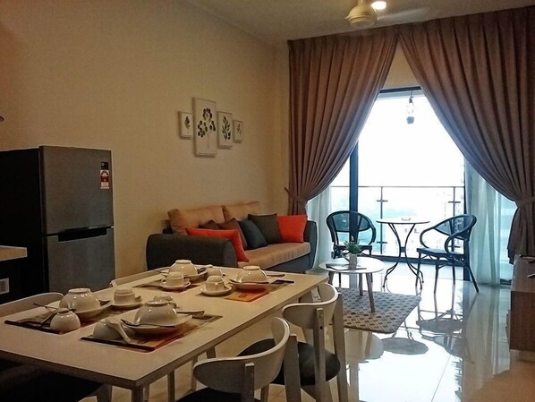 A modern living room with a dining area, a sofa with cushions, a refrigerator, and a balcony with chairs and a table. - Pool View Suites (Johor Bahru)