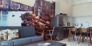 A modern living room with a large cityscape mural, a grey sofa, wooden chairs, a dining table, and a kitchen area with a refrigerator and microwave.