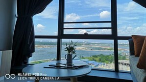 Ein Zimmer mit großem Fenster und Blick auf ein Gewässer sowie entfernte Gebäude. Auf einem kleinen Tisch stehen eine Pflanze und einige Gegenstände.