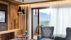 Ein Hotelzimmer mit einem Bett, zwei Sesseln, einem kleinen Tisch und Blick auf Berge durch ein großes Fenster.