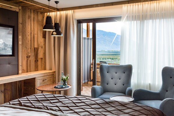 A hotel room with a bed, two armchairs, a small table, and a view of mountains through a large window. - MONDI Hotel Tscherms (Cermes)