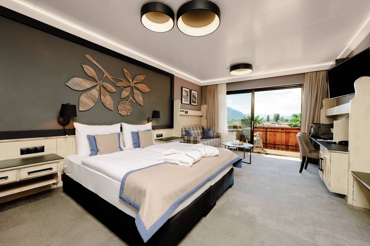 A modern hotel room with a large bed, a desk, a chair, a TV, and a balcony with a view.