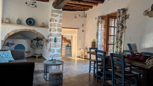 A rustic interior with a stone fireplace, wooden beams, a dining area with blue chairs, and a staircase leading to an upper level.